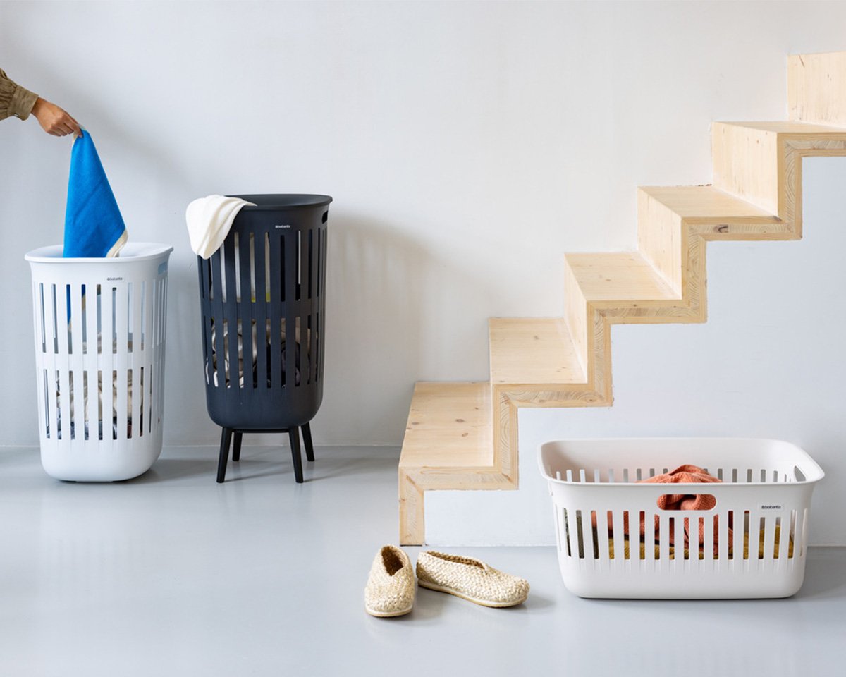 Collect-it wasmand in zwart en wit Drie wasmanden in verschillende kleuren en maten. In een kamer met een grijze vloer en muren met een houten trap. De mand links is wit, 55L; de middelste is zwart, 55L met poten; en de rechter is wit, 40L.